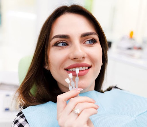 Woman holding clear aligner in dental office setting, smiling at camera.