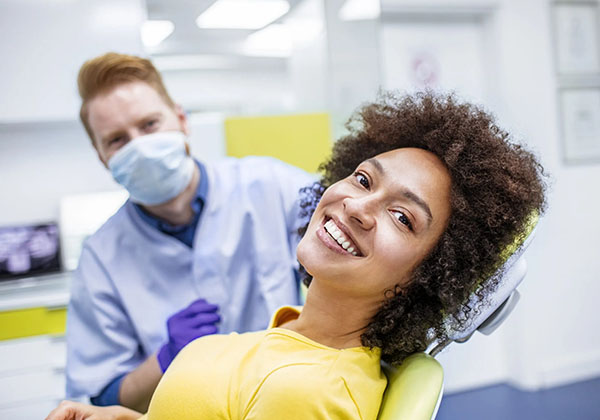 Dental patient in yellow shirt smiling in modern clinic with masked dentist in background.