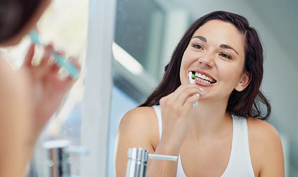 Woman brushing teeth in bathroom mirror, smiling during morning dental routine.